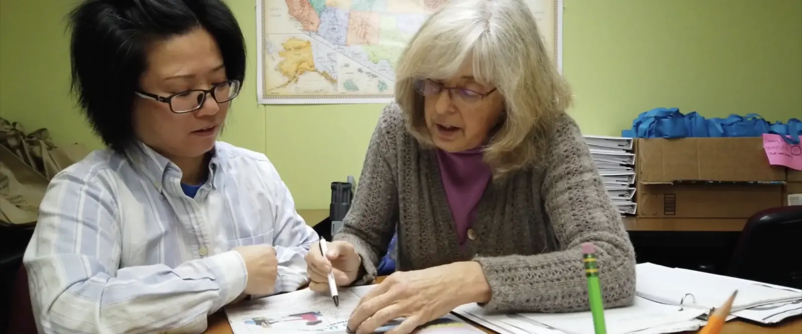A tutor in a purple cardigan points to a workbook while a student with glasses follows along.
