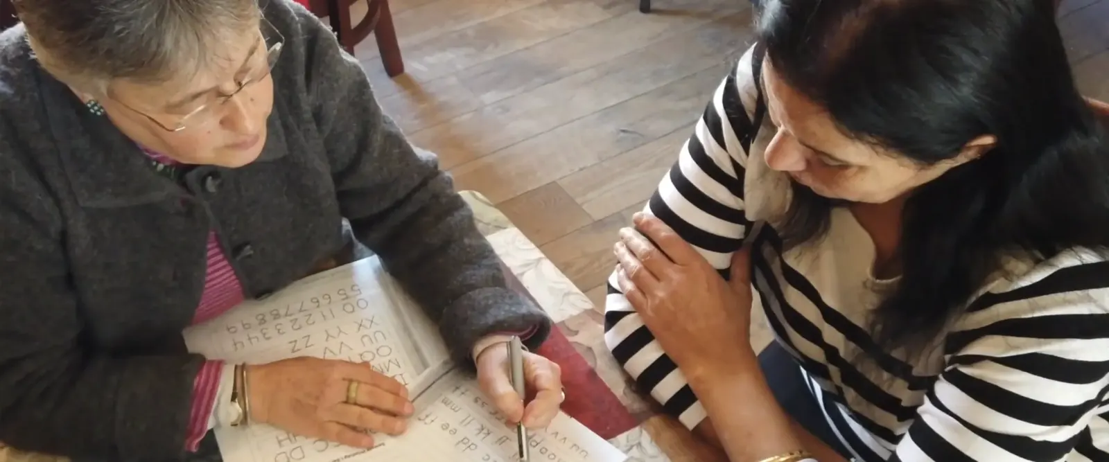 A literacy tutor and a student working together at a wooden table while the student writes in a practice workbook.